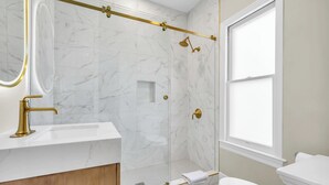 A modern bathroom with a marble shower, a gold showerhead, and a white sink.