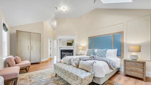 A bedroom with a large bed, a bench, a wardrobe, a fireplace, and a nightstand.