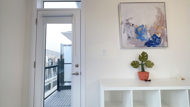 Een witte deur die leidt naar een balkon met uitzicht op gebouwen. Op de muur hangt een lijst met een abstract schilderij en staat een pot met een plant.