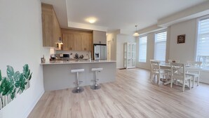 Een moderne keuken met houten kastjes, een wit aanrecht en een ingebouwde koelkast. Er staan twee witte barkrukken bij een eettafel met witte stoelen.
