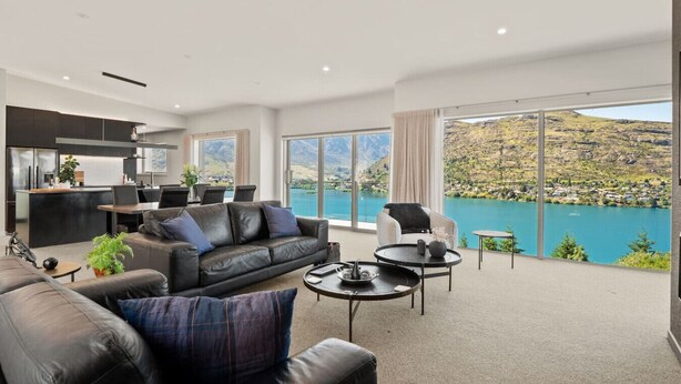 Un salon moderne comprenant un canapé, une table basse et des fauteuils, un coin cuisine et de grandes fenêtres offrant une vue sur un lac et des montagnes.