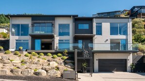 Une maison moderne à deux étages, dotée d’un garage, de grandes fenêtres et d’un jardin aménagé avec des rochers et des arbustes.