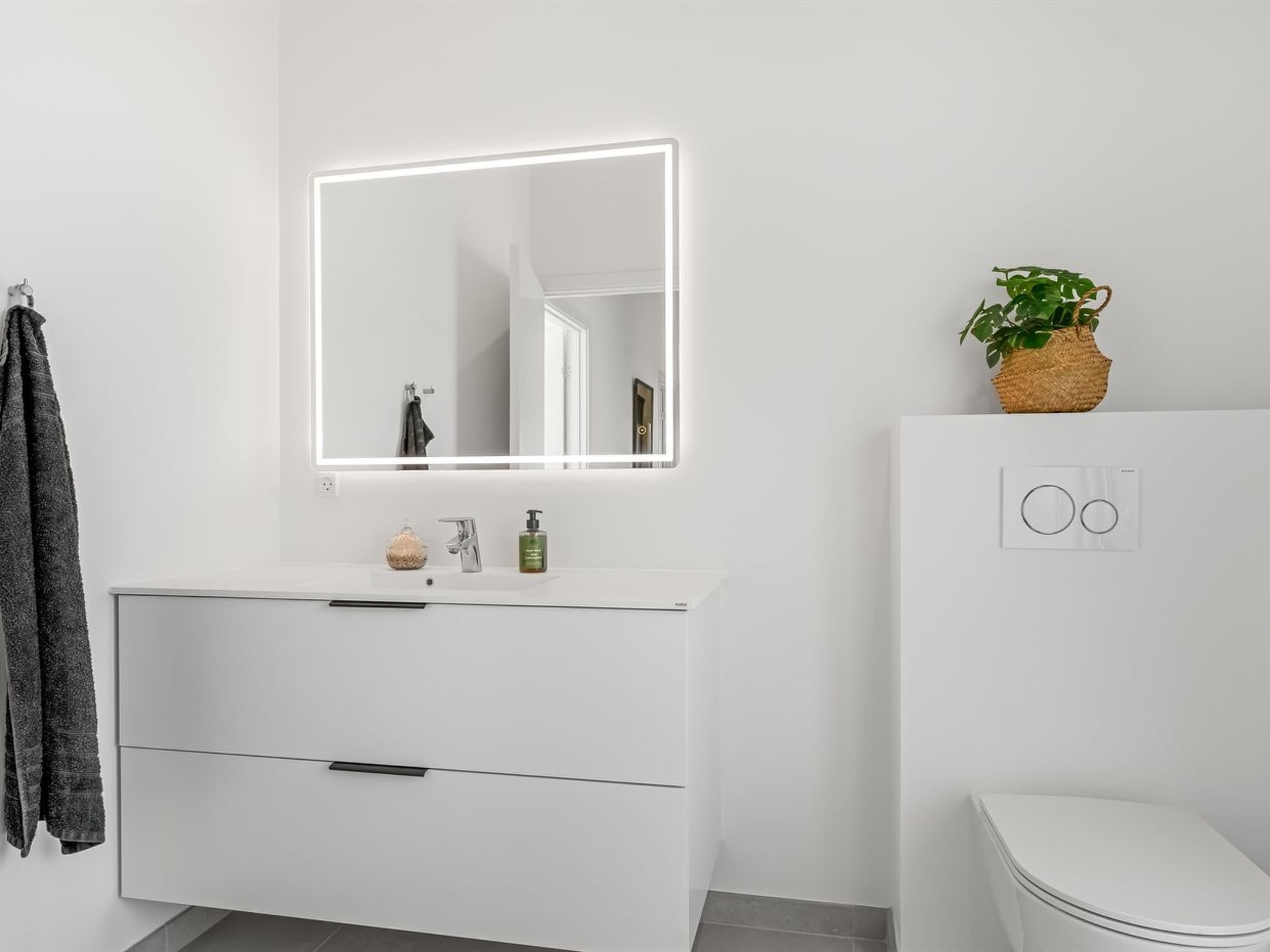 A modern bathroom with a white vanity, a mirror with built-in lighting, a toilet, and a small potted plant.