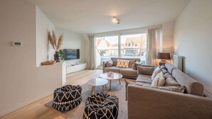 A modern living room with a sofa, two ottomans, a coffee table, and a TV on a wall. - Apartment on Belgian Coast Near Beaches (Knokke-Heist)