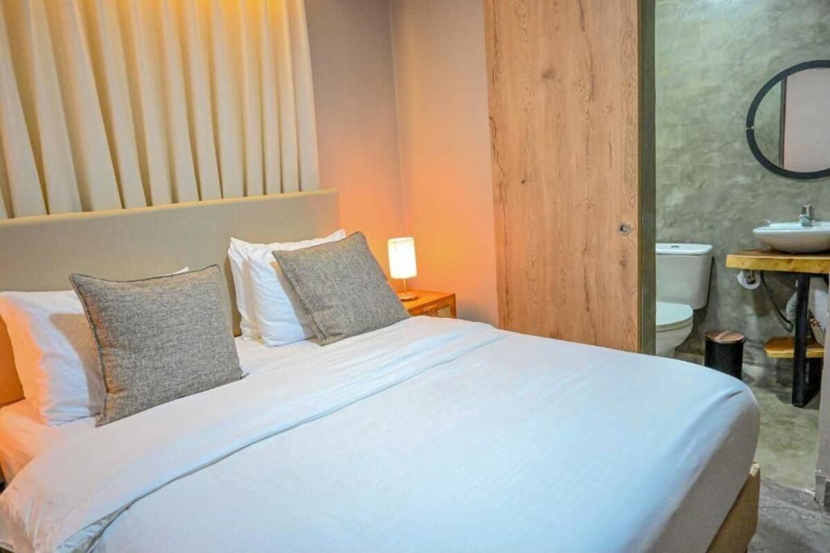 A bedroom with a bed, pillows, a nightstand, a lamp, a wooden wall, and a bathroom with a sink and toilet.