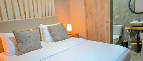 A bedroom with a bed, pillows, a nightstand, a lamp, a wooden wall, and a bathroom with a sink and toilet.