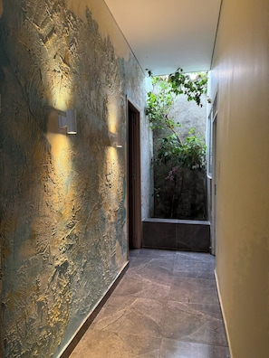 Un pasillo con paredes con textura, piso de baldosas y una puerta que da a una habitación con vistas al verdor.