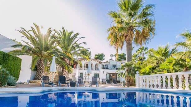 A swimming pool with lounge chairs and umbrellas, surrounded by palm trees and a white fence. - Serene Stay in Benissa (Benissa)