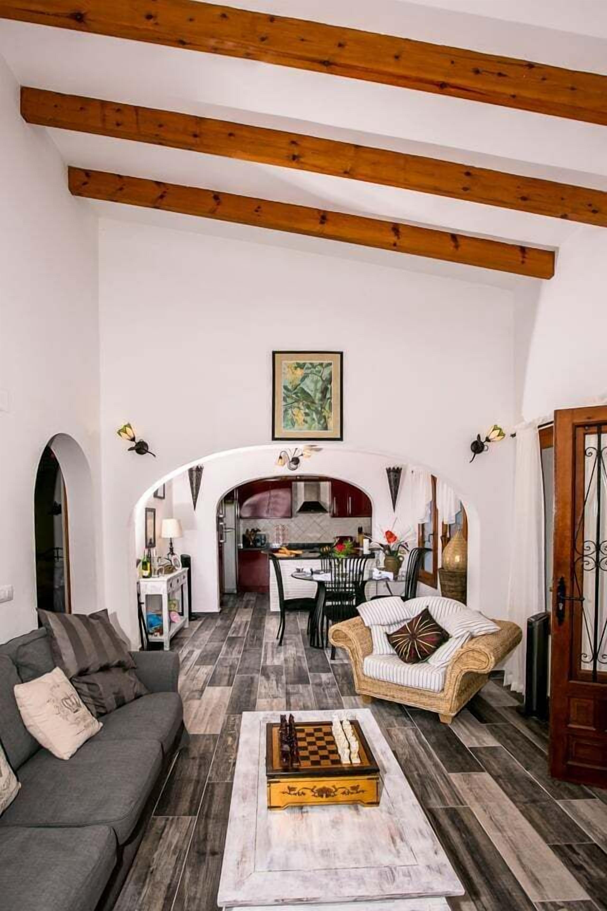 A spacious living room with a wooden ceiling, a sofa, a coffee table with a chess set, and a dining area in the background.