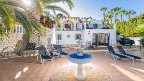 A villa with a white exterior, a stone wall, a tiled patio, lounge chairs, a fountain, and palm trees. - Serene Stay in Benissa (Benissa)