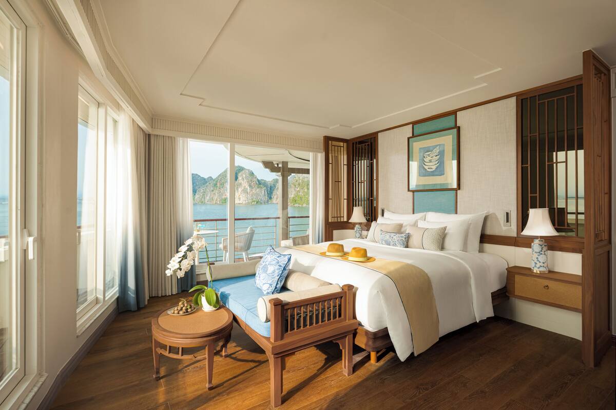 A spacious bedroom with a large bed, a wooden chair, a small table, and a view of the sea through large windows.