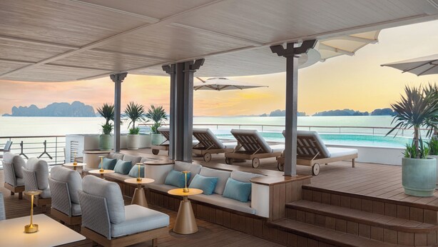 A sunlit outdoor lounge area with seating, tables, and umbrellas, overlooking a calm sea and distant mountains. - Bhaya Soul - Wellness Inclusive Cruise (Tuan Chau)