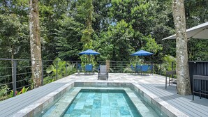 A rectangular pool with a stone border, surrounded by a wooden deck and shaded by two blue umbrellas.