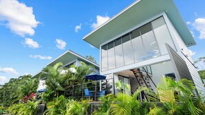 A modern two-story building with large windows, a balcony with chairs and an umbrella, surrounded by lush greenery.