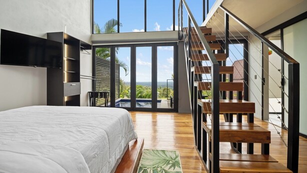 A modern bedroom with a large bed, a wooden staircase, and a view of the outdoors.