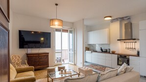 Una sala de estar con sofá, mesa de centro y televisor, una cocina con alacenas blancas y un balcón con vistas.