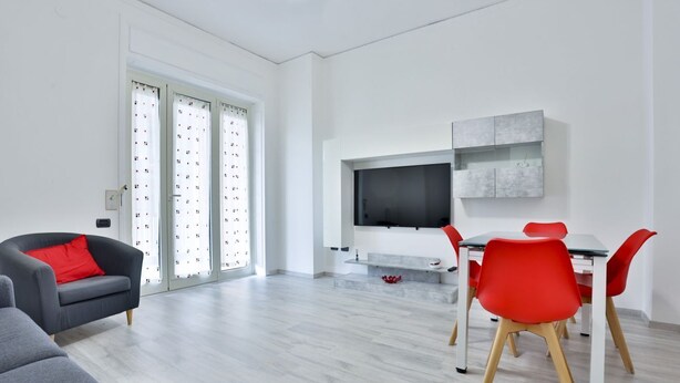 Una sala de estar moderna con un sofá, una mesa de comedor con sillas, un televisor montado en la pared y un ventanal corredero.