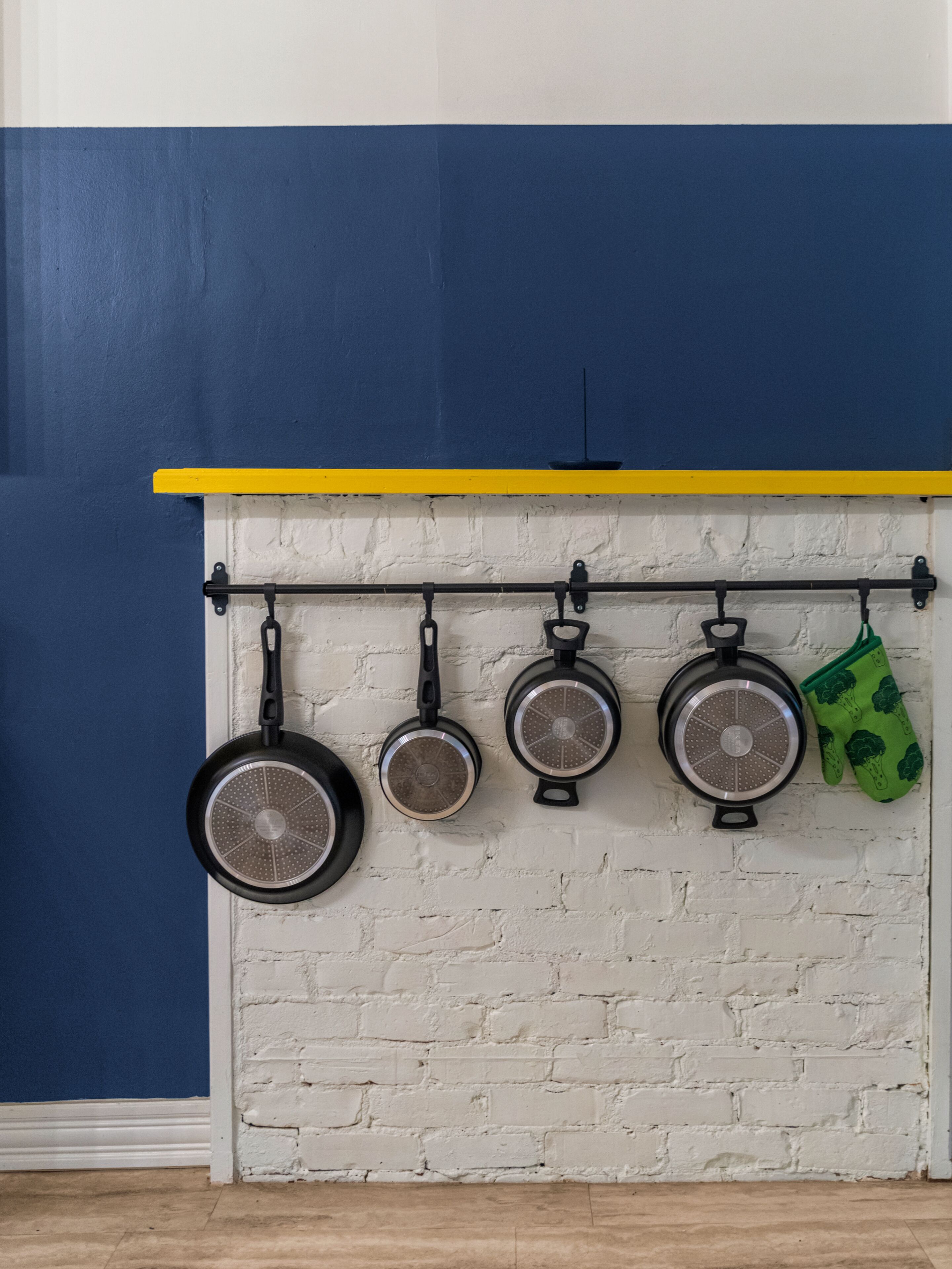 Eine Küche mit weißer Backsteinwand, blauer Akzentwand und einem gelben Regal. Vier Töpfe hängen an einem schwarzen Gestell und ein grüner Ofenhandschuh ist rechts zu sehen.