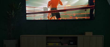 Un televisor mostrando un combate de boxeo, un estante con juegos de mesa y aperitivos sobre una mesa.