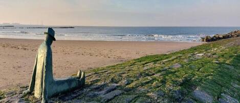 Una estatua de una figura sentada en una playa rocosa, con el océano al fondo.