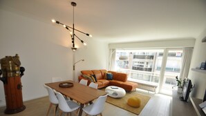A modern living room with a wooden dining table, white chairs, an orange sofa, and a large window offering a city view. - Modern 5th Floor Apartment (Knokke-Heist)