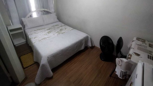 A small, single-bed room with a wooden floor, a white bedspread, a small table with a fan, and a chair. - Azure Flats (Curitiba)