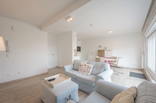 Un salon moderne avec un canapé, un pouf et une petite table sur laquelle repose un plateau. On aperçoit à l’arrière-plan un coin repas avec une table et des chaises.