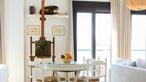 Uma sala de jantar com uma mesa redonda de vidro, cadeiras brancas, uma fruteira e uma pintura em um cavalete.