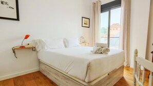 Quarto com piso de madeira, cama com roupa de cama branca, mesa de cabeceira pequena com abajur e janela com cortinas.