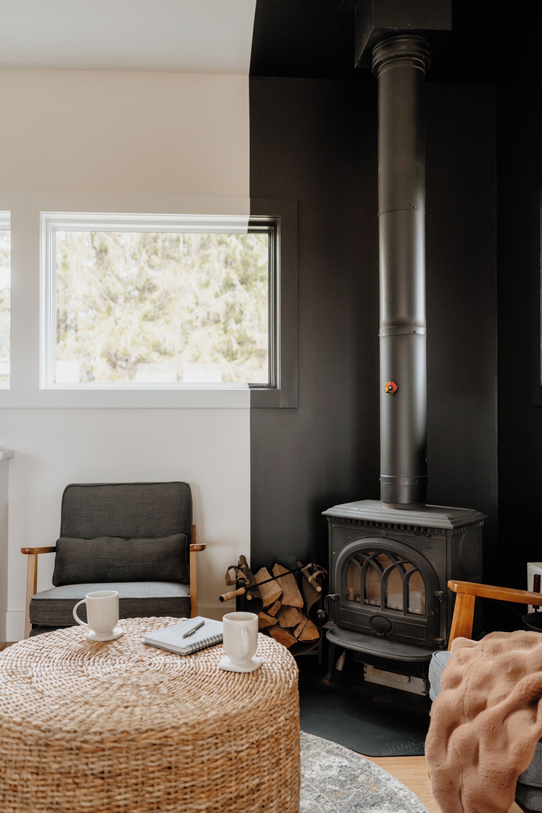 A cozy room with a black wood-burning stove, a comfortable chair, a round woven ottoman, and a large window offering a view of trees.
