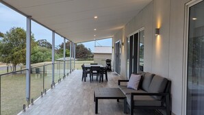 Un patio al aire libre techado con mesa y sillas, un sofá y baranda de cristal.