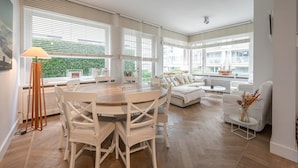 A spacious living area with a round dining table, white furniture, large windows, and a view of greenery outside. - Villa in Knokke Near Alberstrand Beach (Knokke-Heist)