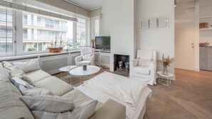 A modern living room with a sofa, a TV, a fireplace, and a view of a building outside. - Villa in Knokke Near Alberstrand Beach (Knokke-Heist)