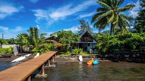 A wooden dock extending over calm water, surrounded by lush greenery and a clear blue sky. - Toahere Beach House - Beachfront - Pool - Pontoon (Papeari)
