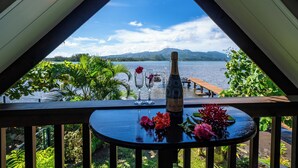 A balcony with a view of a lake and mountains, a bottle, and flowers on a table. - Toahere Beach House - Beachfront - Pool - Pontoon (Papeari)