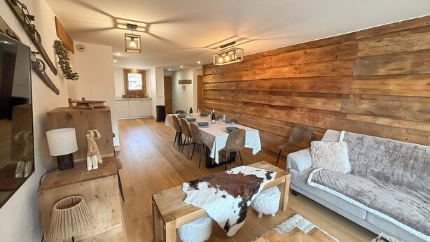 Una sala de estar acogedora con una pared de madera, una zona de comedor con una mesa preparada para cuatro personas y un sofá con un plaid suave.