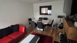 A modern living room with a red sofa, a white coffee table, and a red rug. There is a dining area with a white table and chairs, and a large flat-screen TV mounted on the wall. - Modern Apartment - Quiet Location (Doncaster)