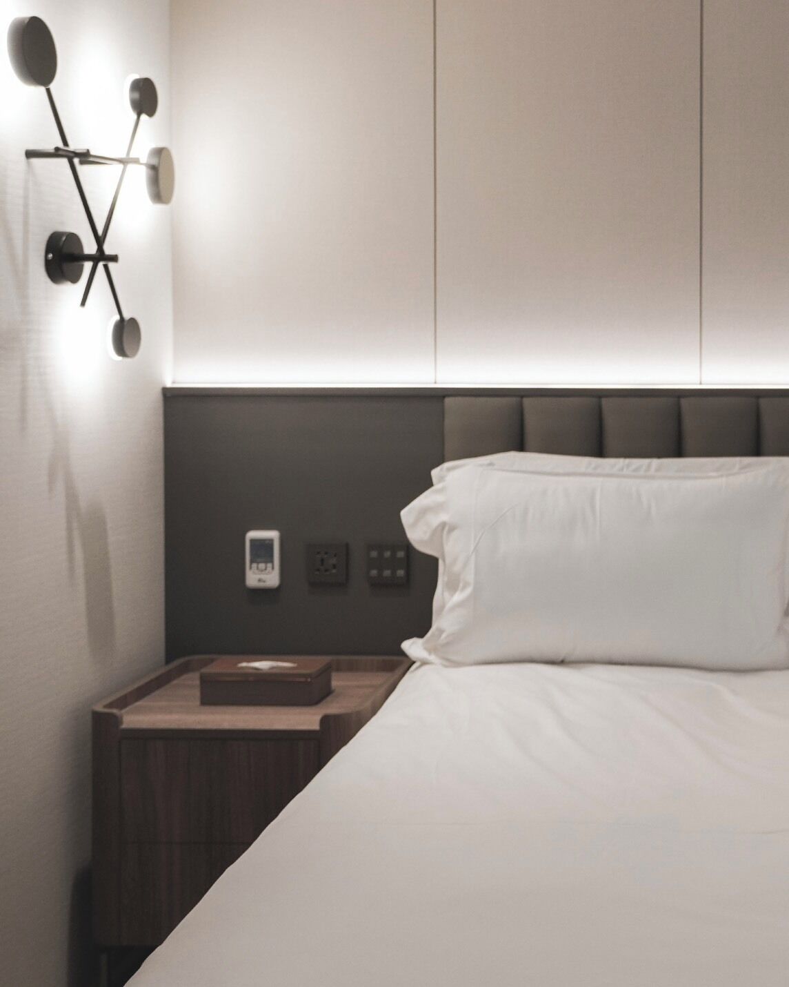 A modern bedroom with a bed, a nightstand, a wall-mounted light fixture, and a control panel.