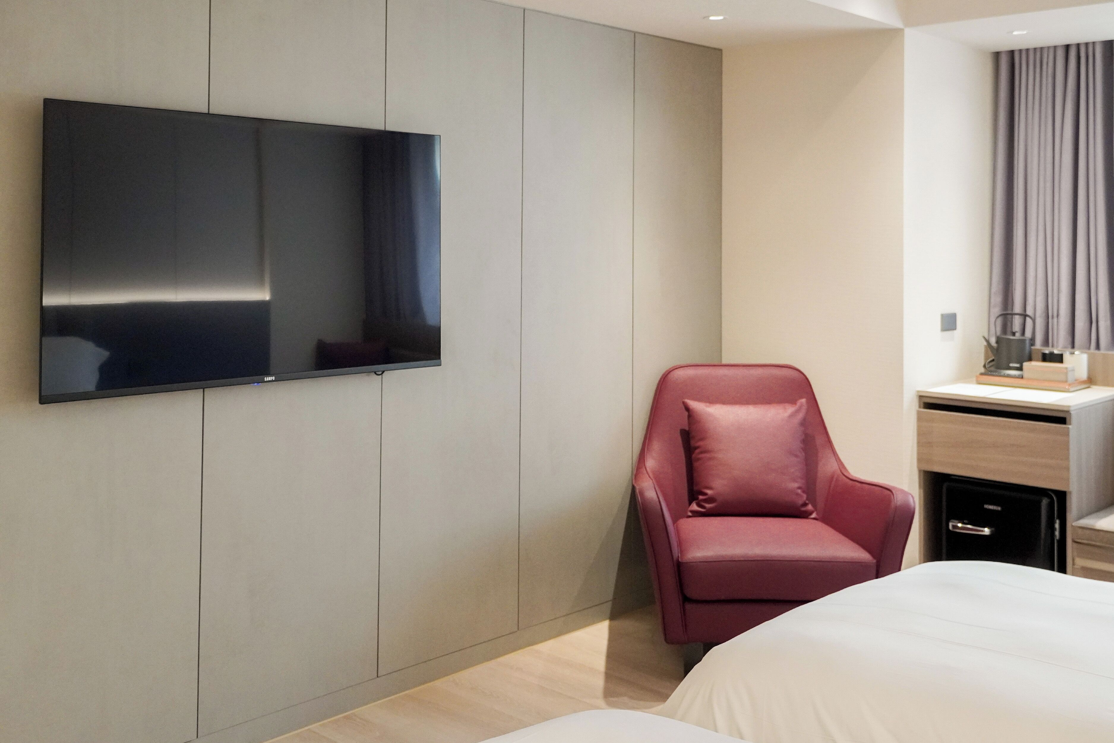A modern hotel room with a large flat-screen TV mounted on the wall, a red leather chair, a bed with white bedding, and a small desk with a computer and a cup.