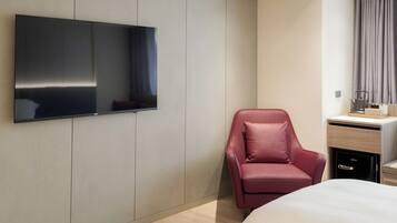 A modern hotel room with a large flat-screen TV mounted on the wall, a red leather chair, a bed with white bedding, and a small desk with a computer and a cup.