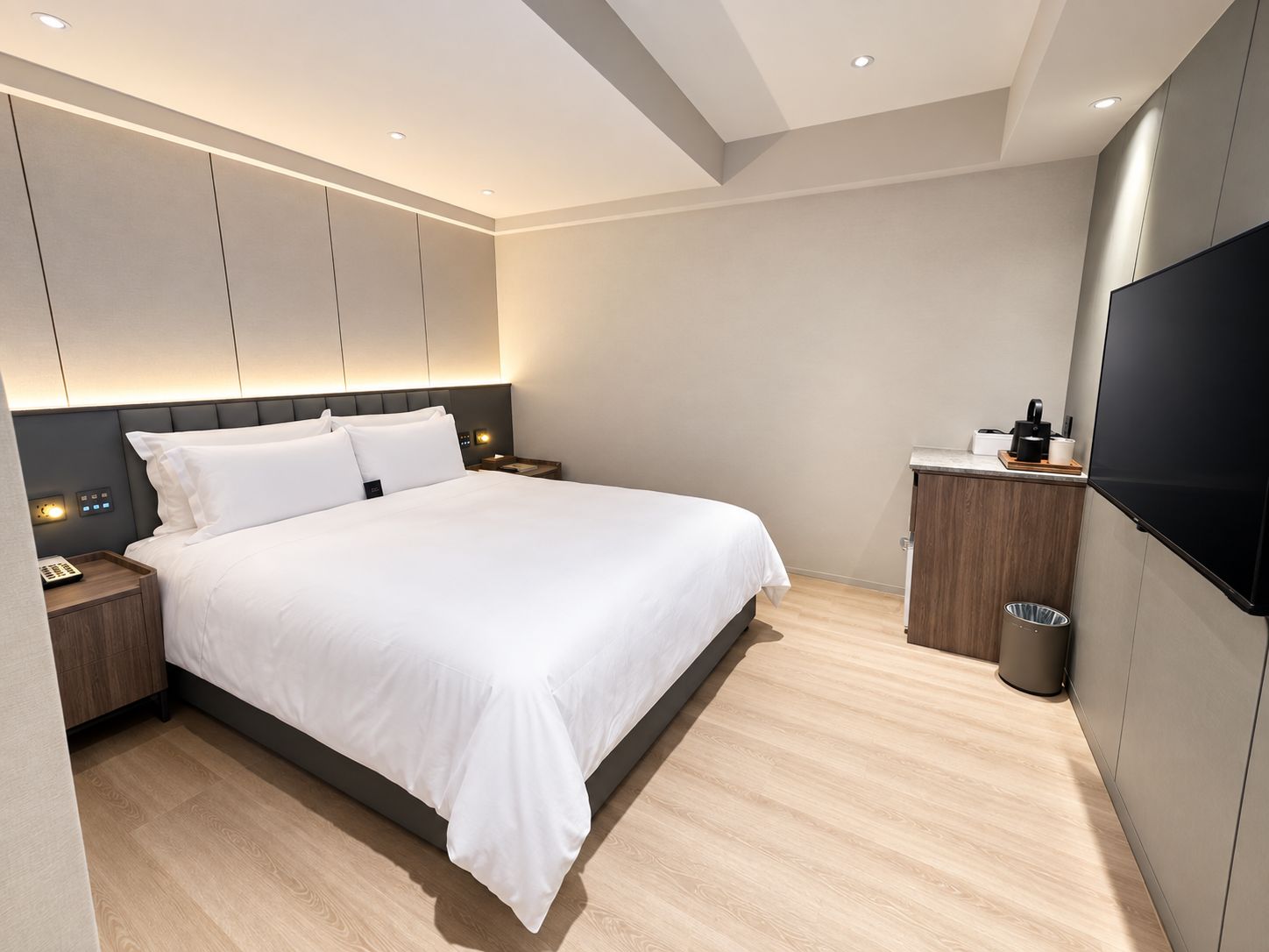A modern hotel room with a large bed, bedside tables, a TV, and a small kitchenette.