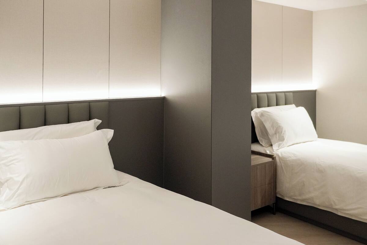 Une chambre d’hôtel moderne avec deux lits, une tête de lit grise et une table de chevet en bois.