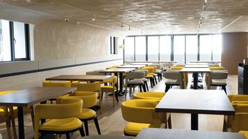 A modern dining area with yellow chairs and wooden tables, large windows, and a curved ceiling.
