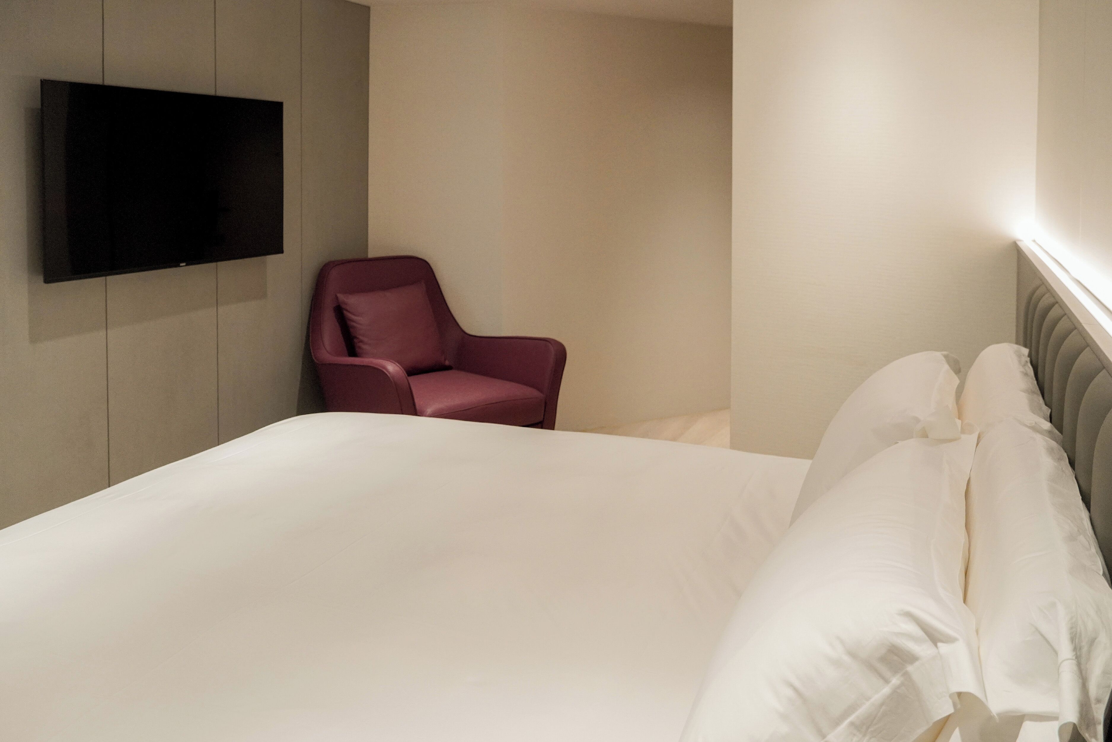 A hotel room with a large bed, a flat-screen TV mounted on the wall, and a single red chair.