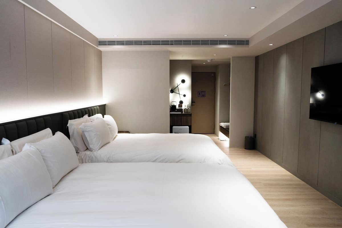 Une chambre d’hôtel moderne dotée de deux grands lits, d’un téléviseur à écran plat fixé au mur et d’un design minimaliste.
