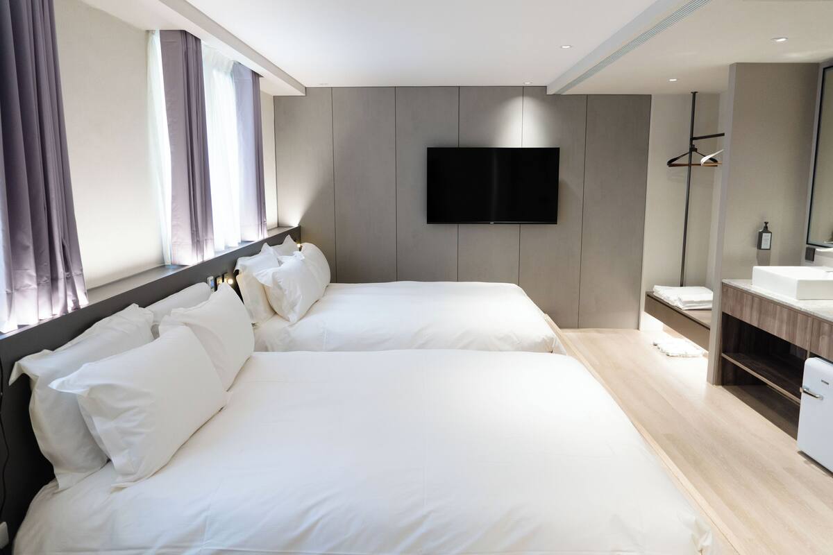 Une chambre d’hôtel moderne avec deux lits, un téléviseur à écran plat, une coiffeuse en bois et une grande fenêtre avec des rideaux.