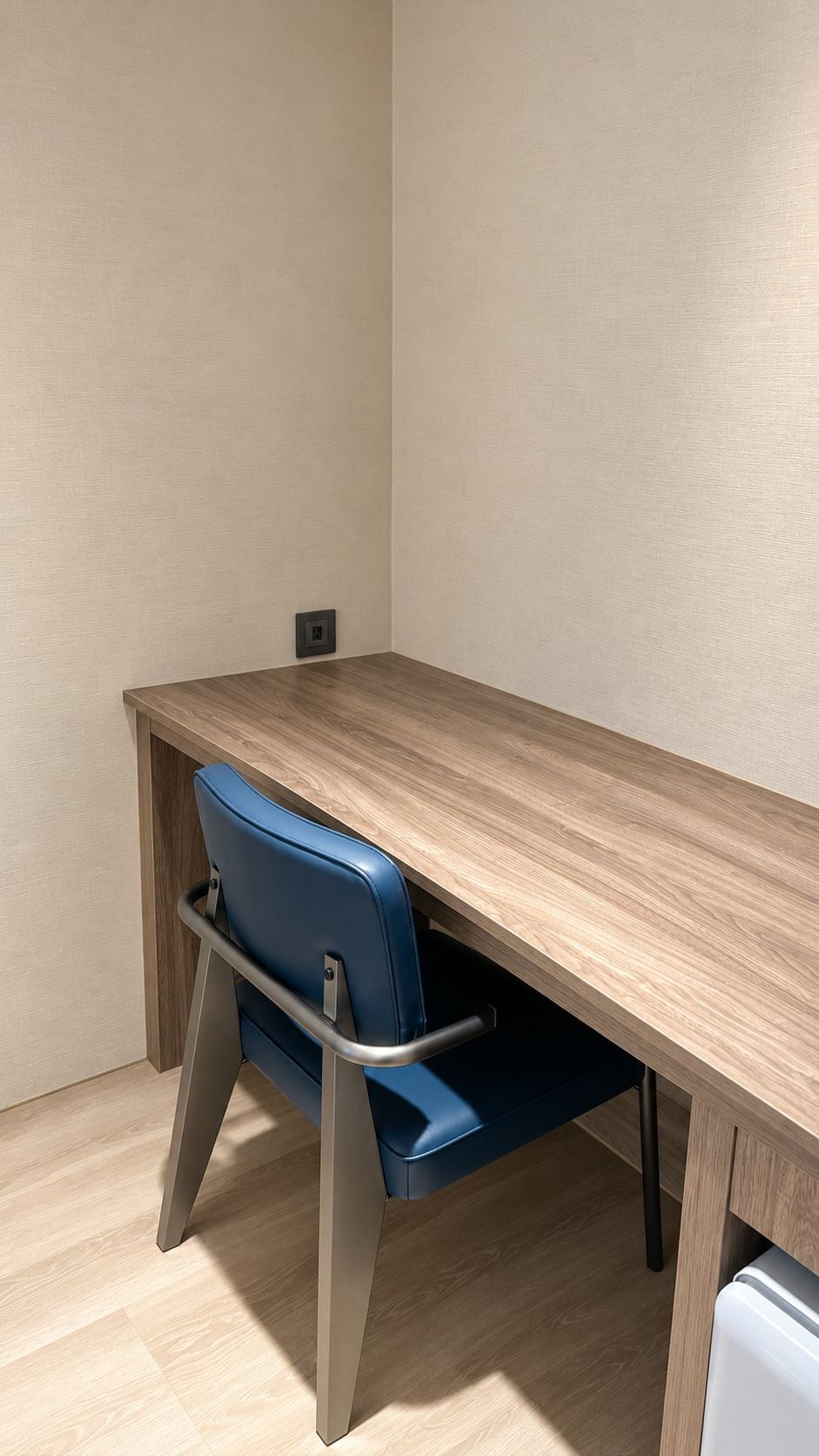 Ein Zimmer mit einem Holztisch, einem blauen Stuhl mit Armlehnen und einem weißen Gerät in der Ecke.