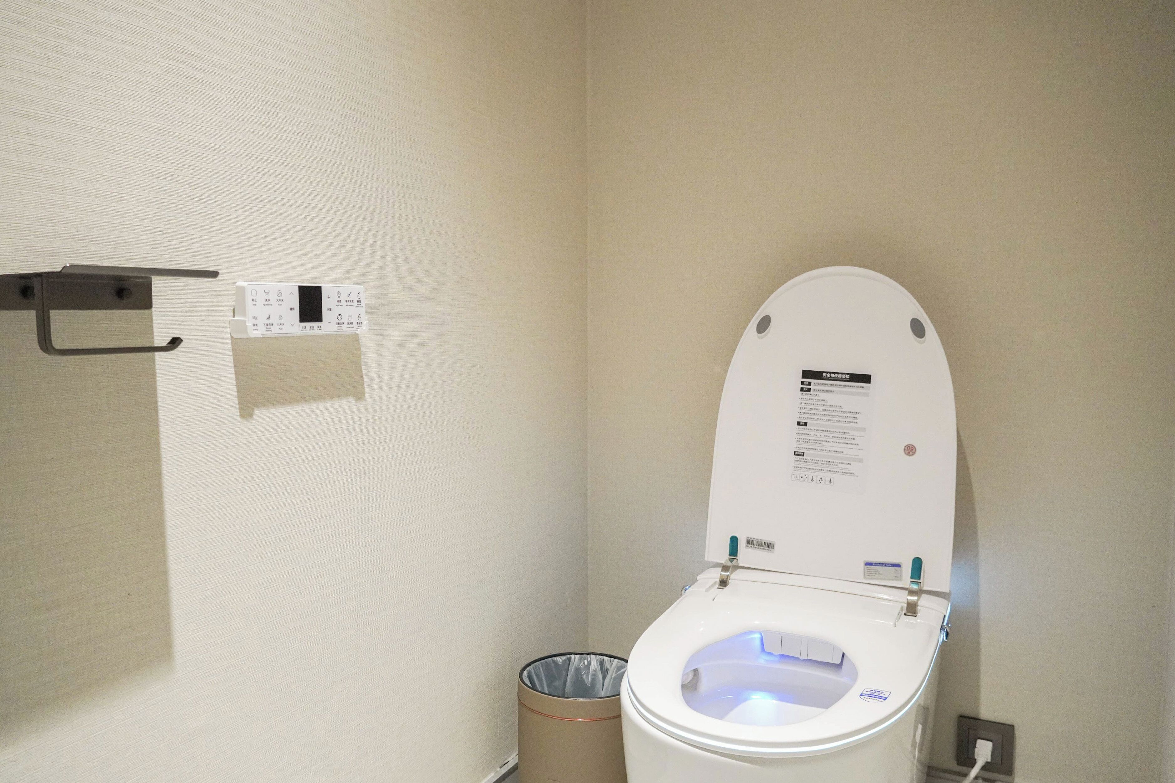 Un baño de hotel estándar con un inodoro blanco, un cesto de basura y un enchufe en la pared.