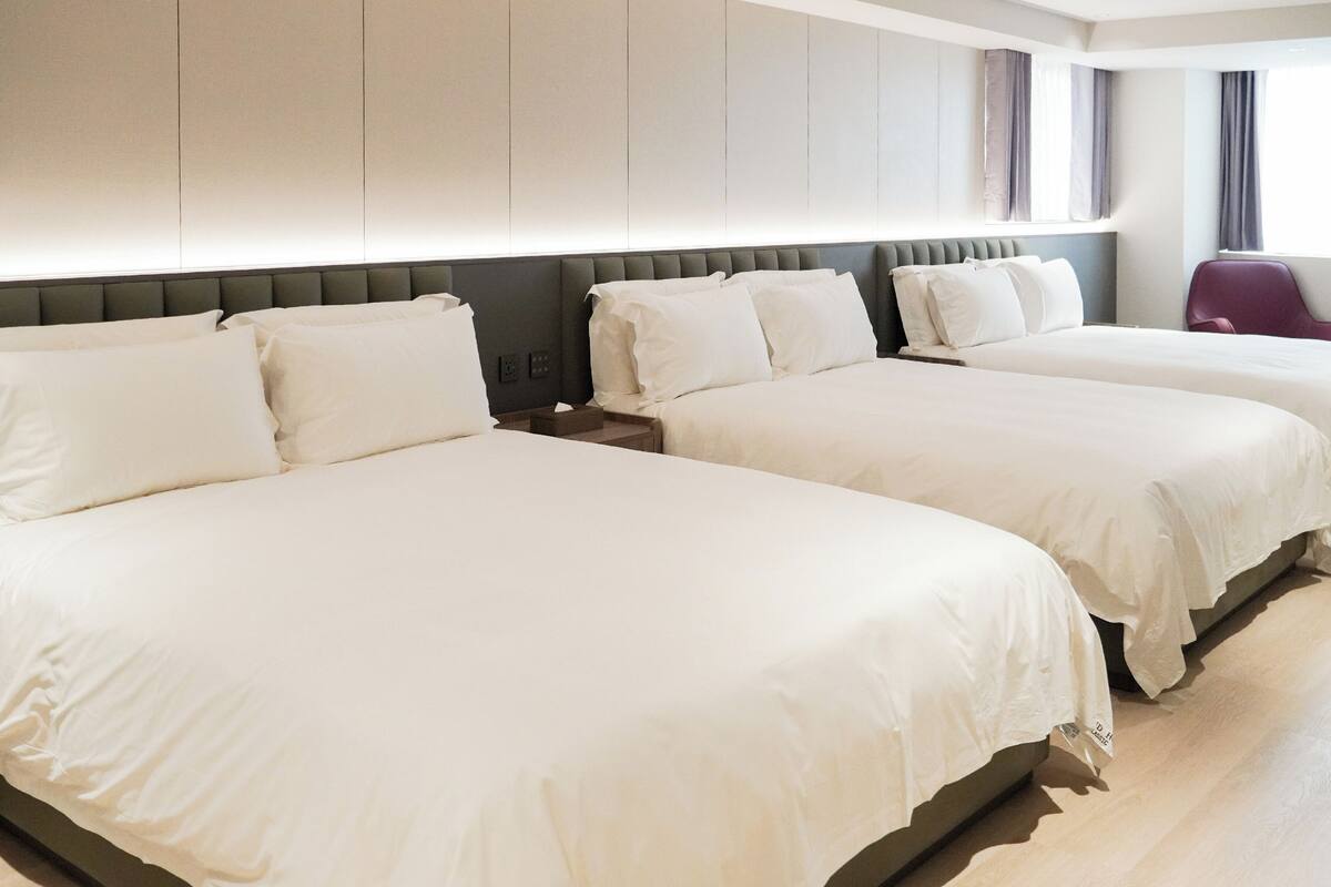 Une chambre d’hôtel avec deux grands lits, une tête de lit grise et une fenêtre avec des rideaux.