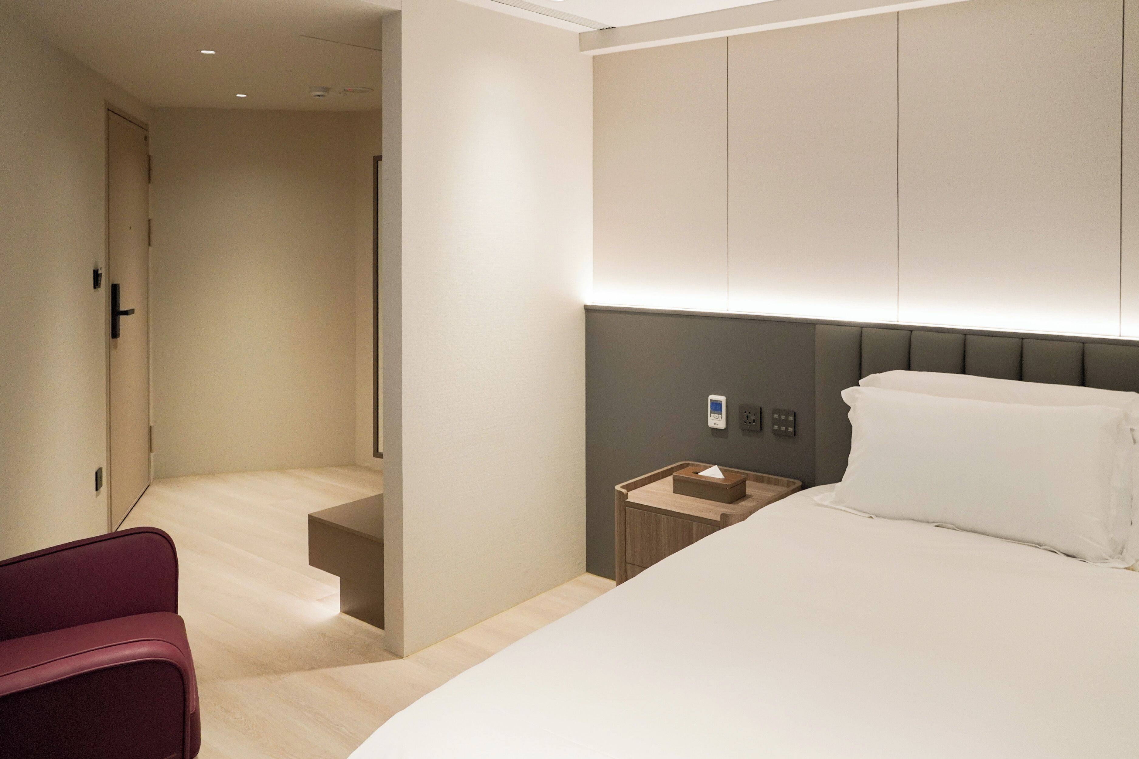 A modern hotel room with a large bed, a bedside table, a nightstand, and a red chair.